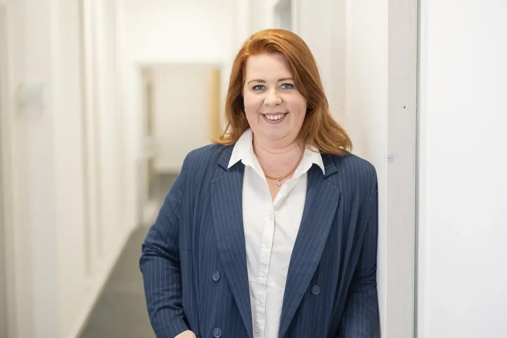 Porträt einer lächelnden Frau mit rotbraunen Haaren, die in weißer Bluse und dunkelblauem Nadelstreifen-Sakko in einem hellen Flur an einer Wand lehnt und in die Kamera schaut.