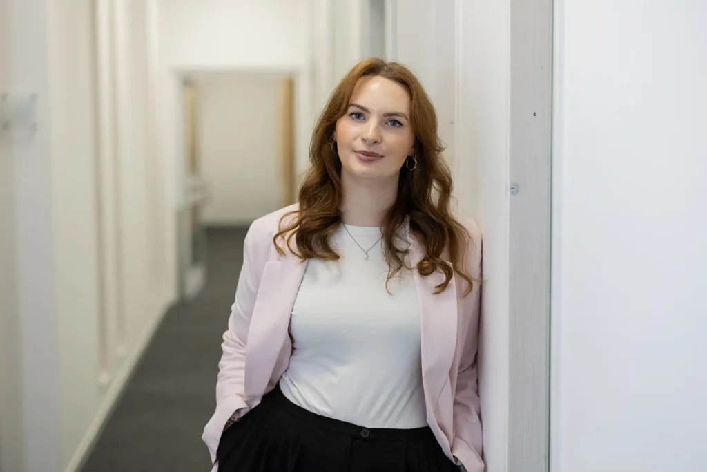 Junge Frau mit schulterlangen, gewellten roten Haaren und silbernen Ohrringen, trägt weißes Shirt und hellrosa Blazer, lehnt mit den Händen in den Taschen lächelnd an einer Wand in einem hellen Büroflur.