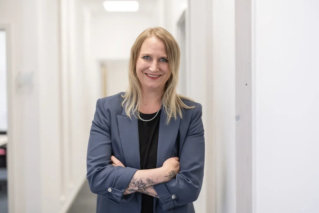 Eine lächelnde Frau mit schulterlangem blondem Haar steht mit verschränkten Armen in einem hellen Büroflur, trägt ein dunkelblaues Sakko über schwarzem Oberteil und zeigt ein florales Unterarmtattoo.