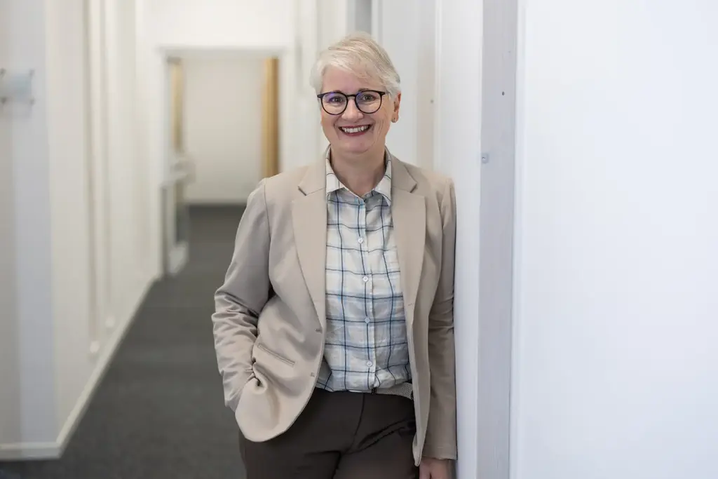 Lächelnde Frau mit kurzem weißem Haar und dunkler Brille, bekleidet mit beigem Blazer und kariertem Hemd, lehnt entspannt an einer Wand in einem hellen Büroflur.