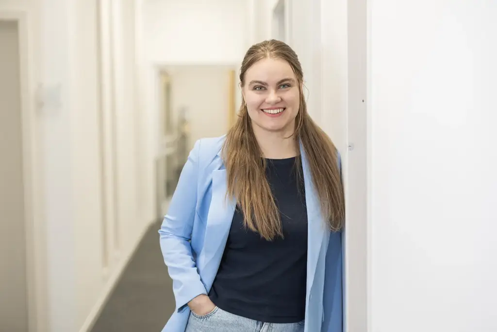 Eine lächelnde junge Frau mit langen braunen Haaren lehnt in einem hellen Büroflur an einer Wand, trägt ein hellblaues Sakko über einem dunklen Oberteil und hat eine Hand in der Hosentasche.