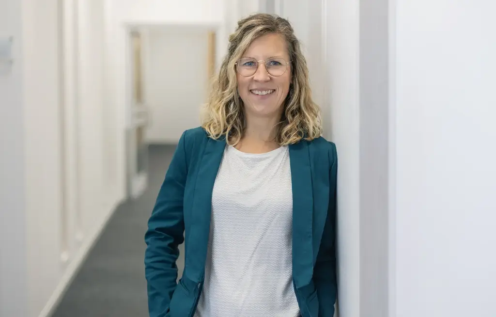 Lächelnde Frau mit lockigem blondem Haar und runder Brille, trägt einen türkisfarbenen Blazer über weißer Bluse und lehnt in einem hellen Büroflur an einer Wand.