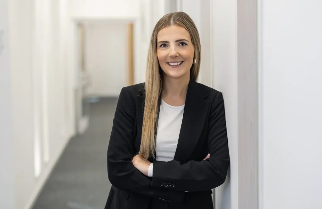 Lächelnde Frau mit langem, glattem blondem Haar und Creolen, in schwarzem Blazer und weißem Shirt, lehnt mit verschränkten Armen an einer weißen Wand in einem hellen Büroflur.