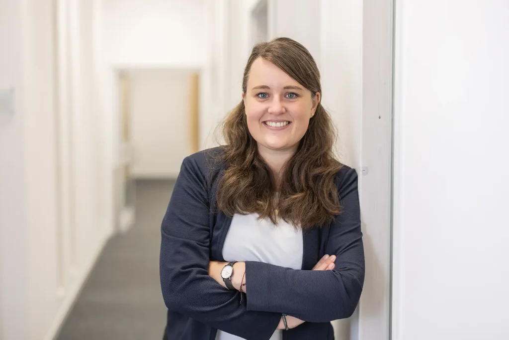 Lächelnde Frau mit langen braunen Haaren und blauem Blazer steht mit verschränkten Armen in einem hellen Büroflur.