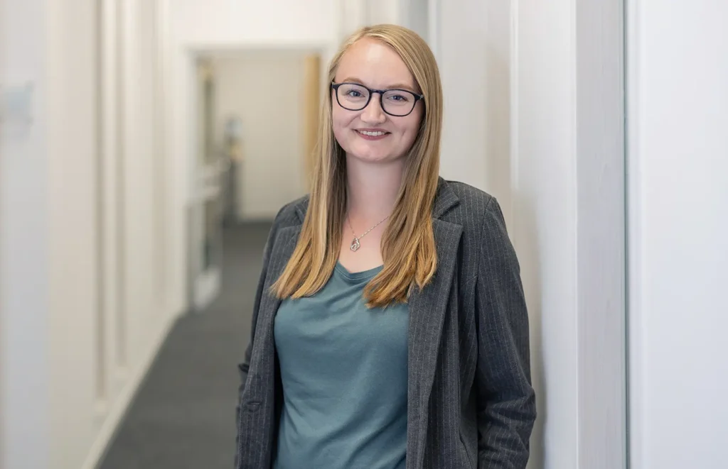 Junge Frau mit langen blonden Haaren und dunkler Brille lehnt lächelnd in einem hellen Büroflur an einer weißen Wand und trägt einen grauen Nadelstreifenblazer, grünes Shirt und eine Herz-Kette.
