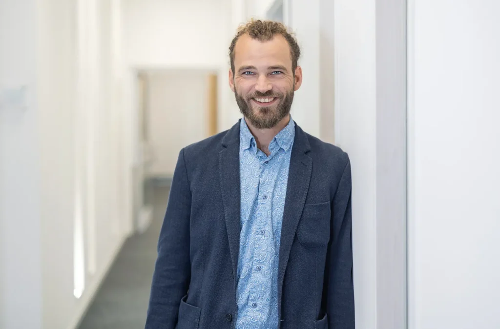 Lächelnder Mann mit Vollbart und zurückgebundenem Haar, in dunkelblauem Sakko und gemustertem hellblauen Hemd, steht in einem hellen Büroflur und lehnt leicht an einer Wand.