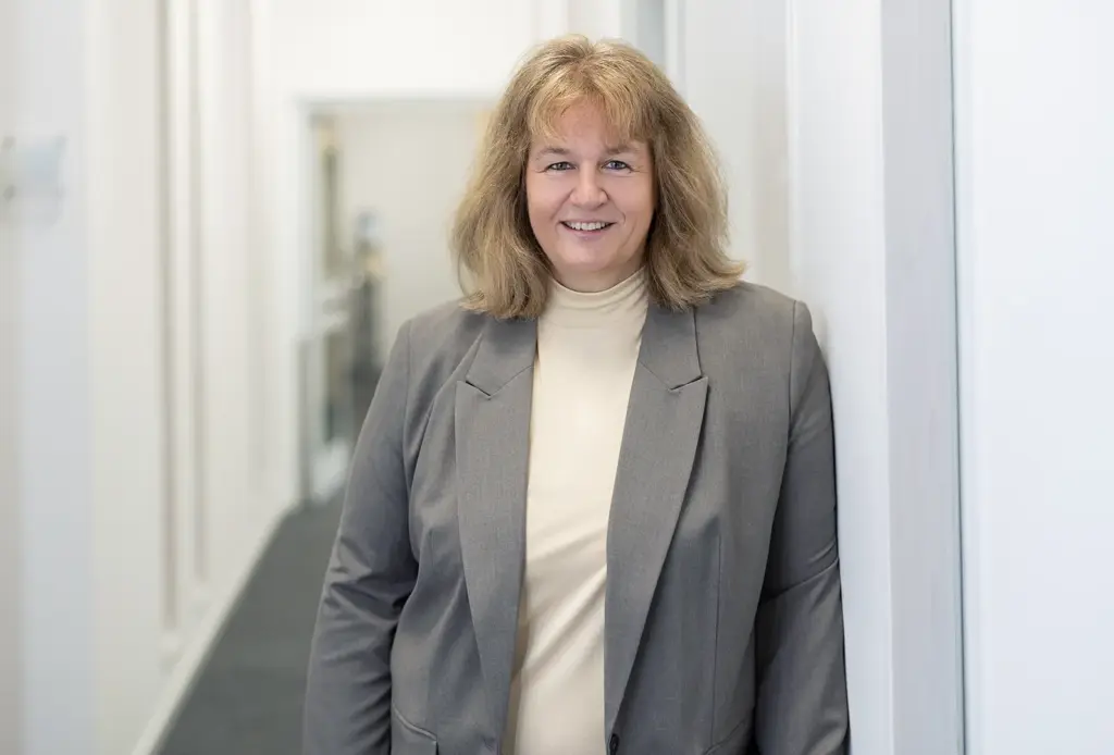 Lächelnde Frau mittleren Alters mit schulterlangem, hellblondem Haar, in hellem Rollkragenpullover und grauem Blazer, die in einem hellen Büroflur an einer Wand lehnt.