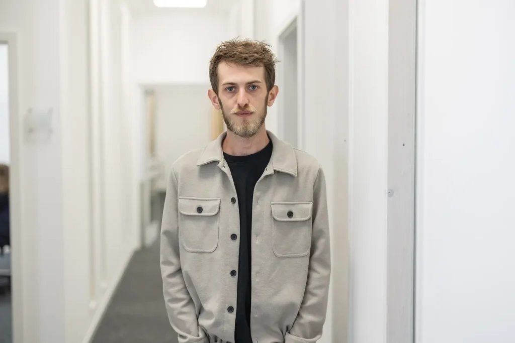 Junger Mann mit kurzem, zerzaustem Haar und Dreitagebart in einer beigen Jacke über schwarzem T‑Shirt, mit den Händen in den Taschen, steht in einem hellen, schmalen Flur und blickt neutral in die Kamera.