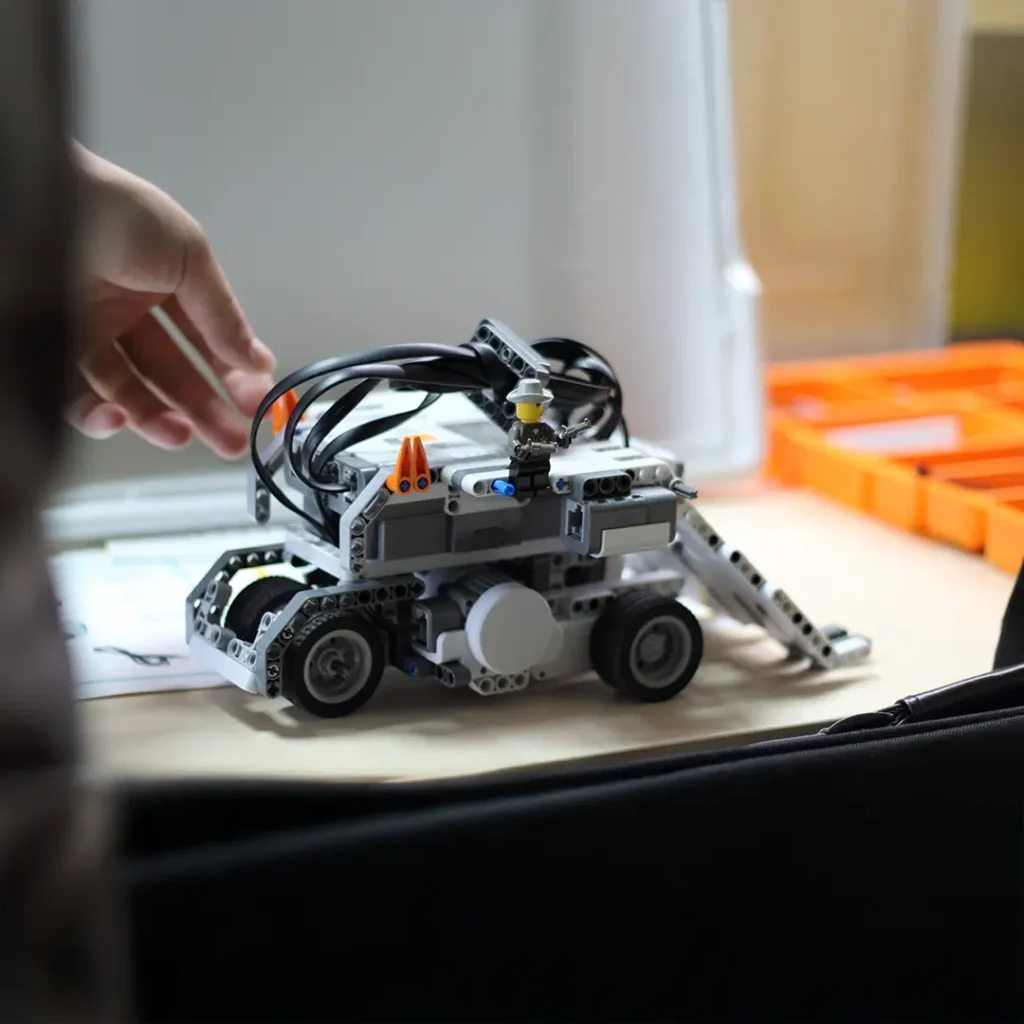 Auf einem Tisch steht ein grauer Lego‑Technic‑Roboter mit Kabeln und Rädern, auf dem eine kleine Minifigur steht; im Hintergrund ist eine orange Aufbewahrungsbox und eine Hand, die nach dem Modell greift.