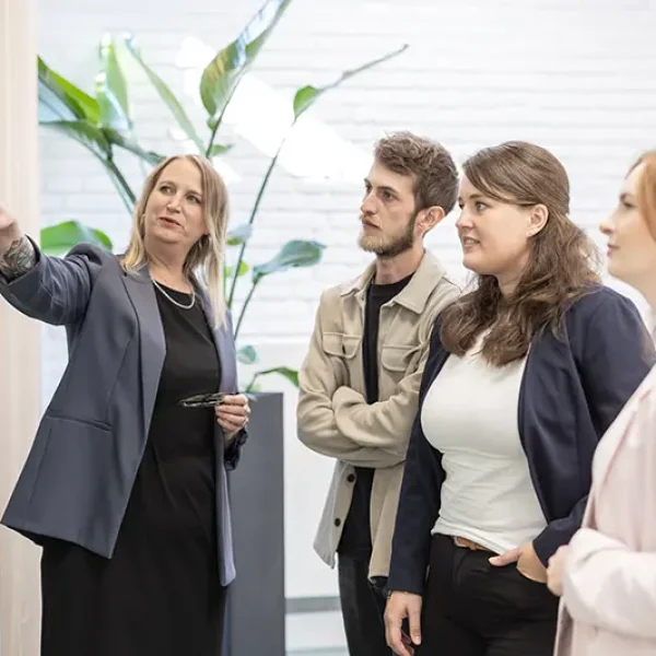 Vier Personen in einem modernen Büro betrachten eine Wandpräsentation; eine Frau links im Blazer zeigt mit ausgestrecktem Arm darauf, die anderen drei (ein Mann und zwei Frauen) blicken aufmerksam.