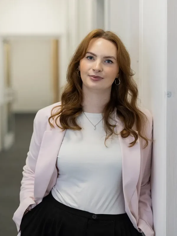 Junge Frau mit schulterlangen, gewellten roten Haaren und silbernen Ohrringen, trägt weißes Shirt und hellrosa Blazer, lehnt mit den Händen in den Taschen lächelnd an einer Wand in einem hellen Büroflur.