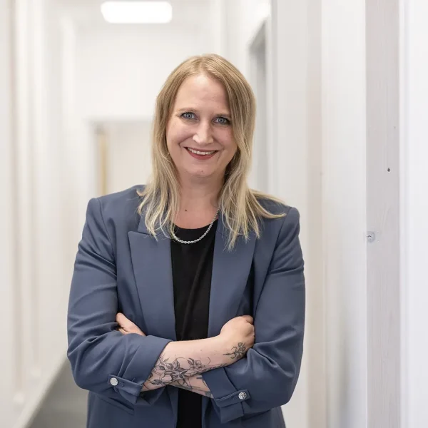 Eine lächelnde Frau mit schulterlangem blondem Haar steht mit verschränkten Armen in einem hellen Büroflur, trägt ein dunkelblaues Sakko über schwarzem Oberteil und zeigt ein florales Unterarmtattoo.