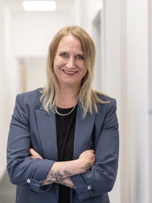 Eine lächelnde Frau mit schulterlangem blondem Haar steht mit verschränkten Armen in einem hellen Büroflur, trägt ein dunkelblaues Sakko über schwarzem Oberteil und zeigt ein florales Unterarmtattoo.
