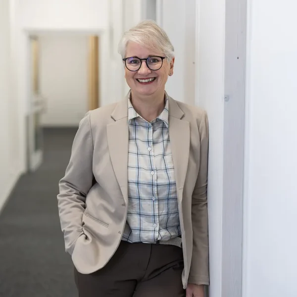 Lächelnde Frau mit kurzem weißem Haar und dunkler Brille, bekleidet mit beigem Blazer und kariertem Hemd, lehnt entspannt an einer Wand in einem hellen Büroflur.
