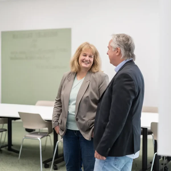 Zwei lächelnde Personen — eine Frau mit hellem Blazer und Jeans und ein Mann mit dunklem Sakko und Jeans — unterhalten sich in einem hellen Besprechungsraum mit Tischen, Stühlen und einer Wandtafel im Hintergrund.