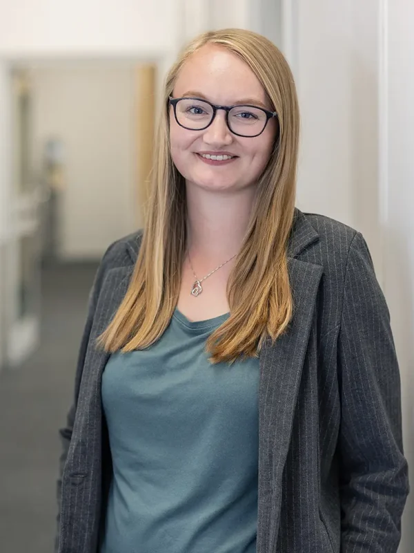 Junge Frau mit langen blonden Haaren und dunkler Brille lehnt lächelnd in einem hellen Büroflur an einer weißen Wand und trägt einen grauen Nadelstreifenblazer, grünes Shirt und eine Herz-Kette.