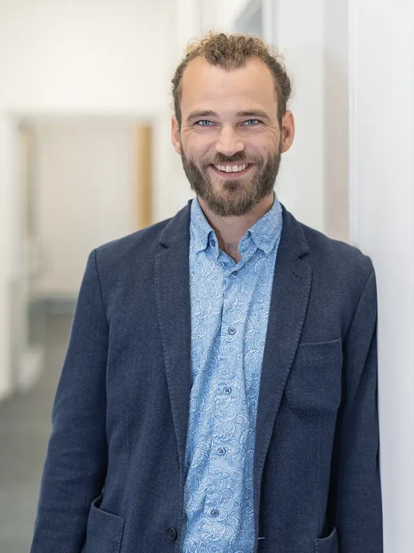 Lächelnder Mann mit Vollbart und zurückgebundenem Haar, in dunkelblauem Sakko und gemustertem hellblauen Hemd, steht in einem hellen Büroflur und lehnt leicht an einer Wand.