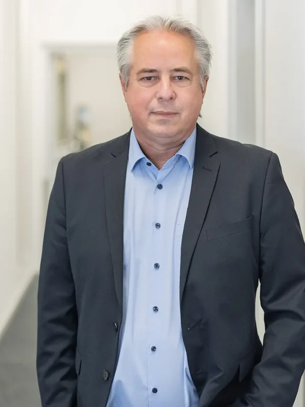 Mann mittleren Alters mit grauem Haar und hellblauem Hemd trägt dunklen Anzug und steht mit nach vorn gerichteter Miene in einem hellen, schmalen Flur.