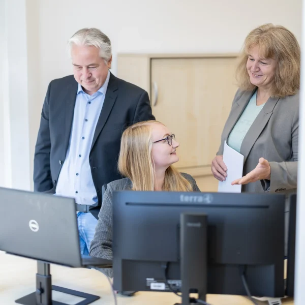 Junge Frau sitzt an einem Schreibtisch vor zwei Monitoren und lächelt, während zwei ältere Kolleg:innen neben ihr stehen und mit Papieren in der Hand ein freundliches Gespräch führen.