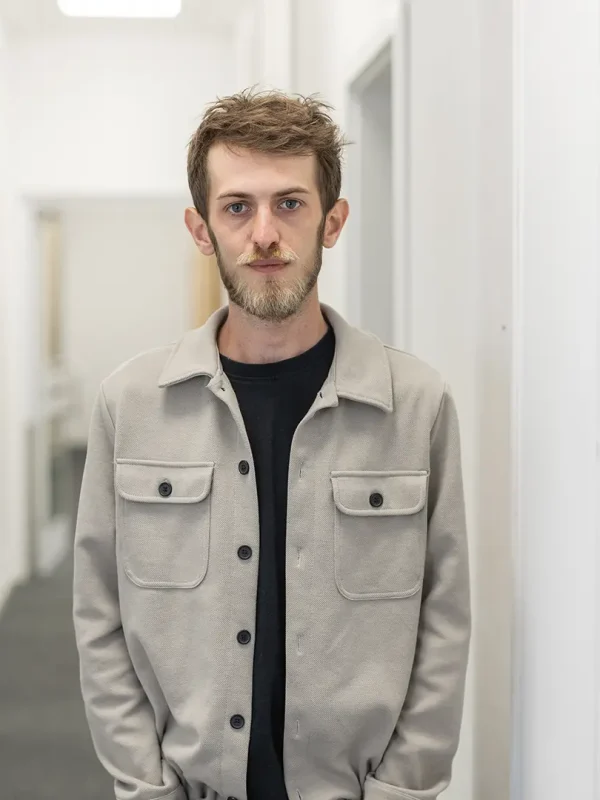 Junger Mann mit kurzem, zerzaustem Haar und Dreitagebart in einer beigen Jacke über schwarzem T‑Shirt, mit den Händen in den Taschen, steht in einem hellen, schmalen Flur und blickt neutral in die Kamera.