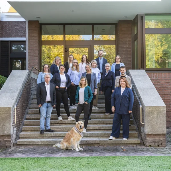 Gruppenfoto von etwa fünfzehn Erwachsenen in Business‑Casual, die auf einer Außentreppe vor einem Backsteingebäude mit großen Fenstern stehen, in mehreren Reihen zur Kamera lächeln, mit einem sitzenden Golden Retriever vorn auf der Treppe und herbstlich gefärbten Bäumen, die sich in den Fenstern spiegeln.