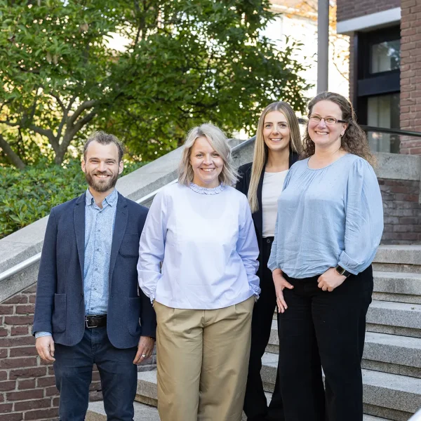 Vier lächelnde Erwachsene (drei Frauen, ein Mann) stehen auf einer Außentreppe vor einem Backsteingebäude und Büschen; der Mann trägt ein Sakko, die Frauen ein weißes Oberteil, ein schwarzes Blazer-Outfit und eine hellblaue Bluse mit Brille.