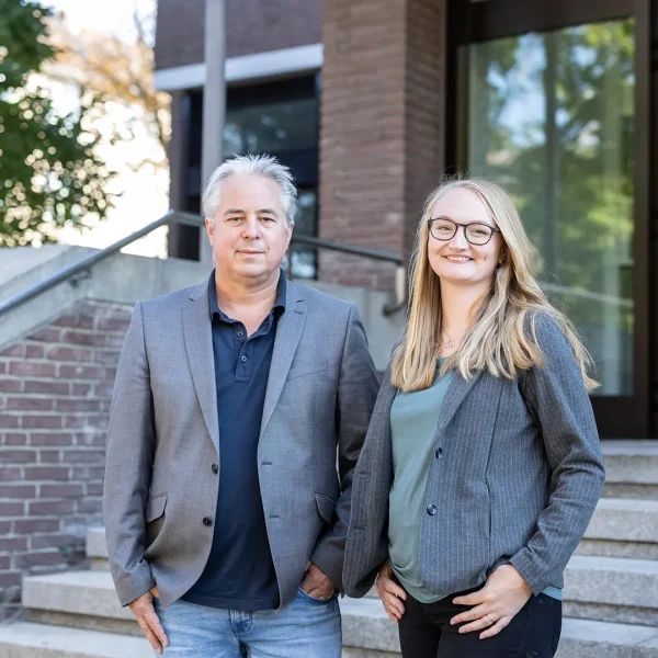 Ein Mann mit grauen Haaren und eine Frau mit langen blonden Haaren und Brille stehen in lässigen Blazern auf einer Außentreppe vor einem Backsteingebäude mit Glastür, die Frau lächelt.
