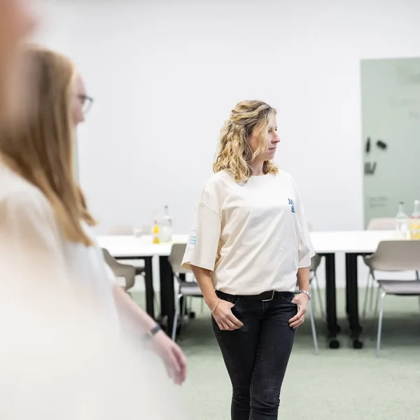 Frau steht in einem hellen Besprechungsraum in lockerem hellen T‑Shirt und dunkler Jeans mit den Händen in den Taschen und schaut zur Seite; im Vordergrund unscharfe Personen, im Hintergrund Tische, Stühle und Flaschen vor einer Glaswand.